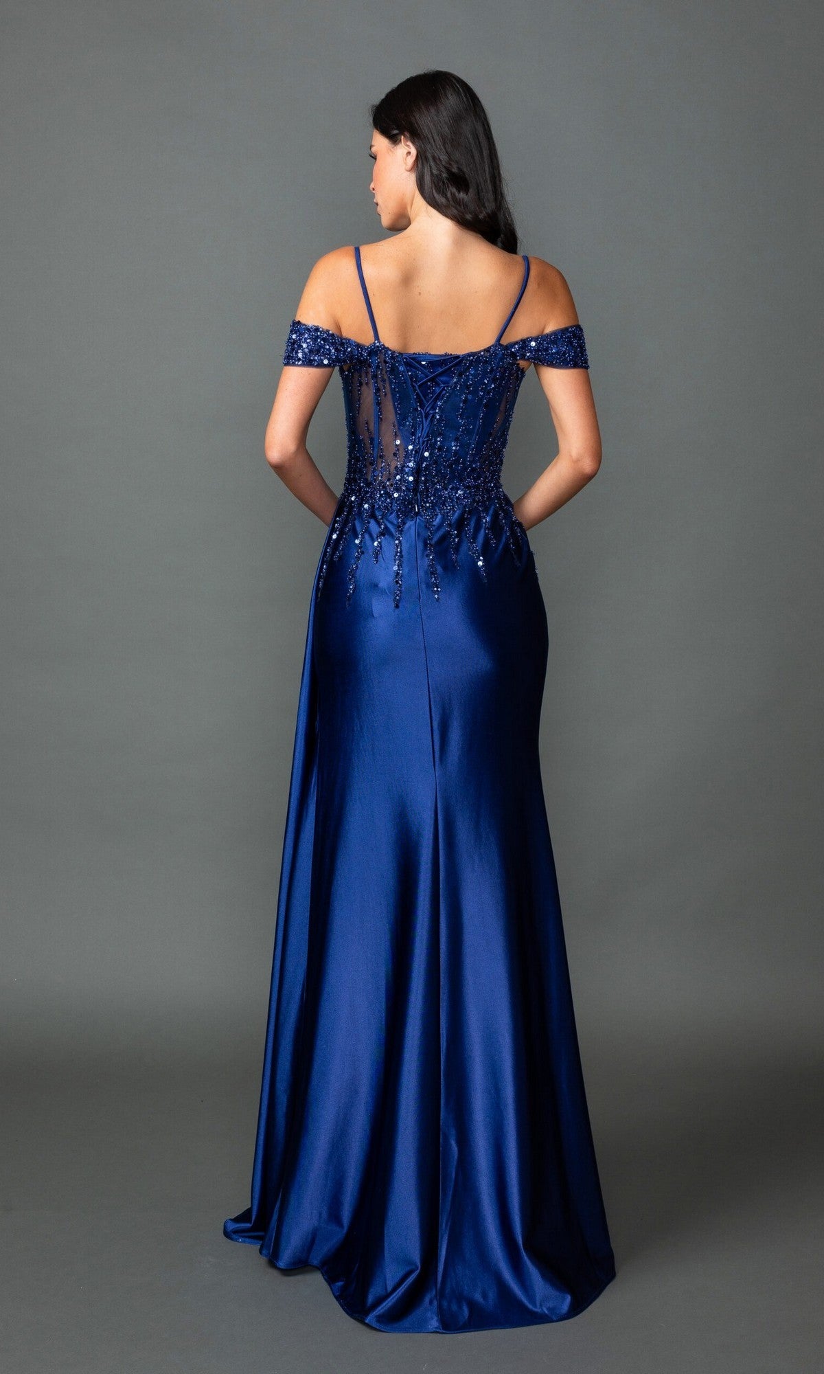 A woman faces away against a plain gray background, wearing the Dancing Queen 4551 long prom dress—an off-the-shoulder, floor-length royal blue gown with sequin-embellished bodice.